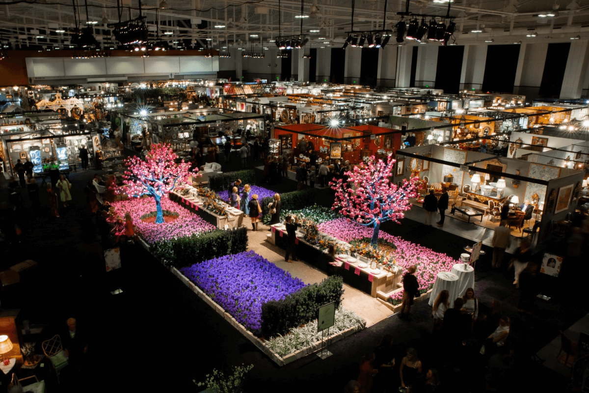 Large indoor exhibition hall with floral displays, including pink artificial trees and purple flowers, surrounded by vendor booths and visitors exploring this vibrant setting for southern events in January.