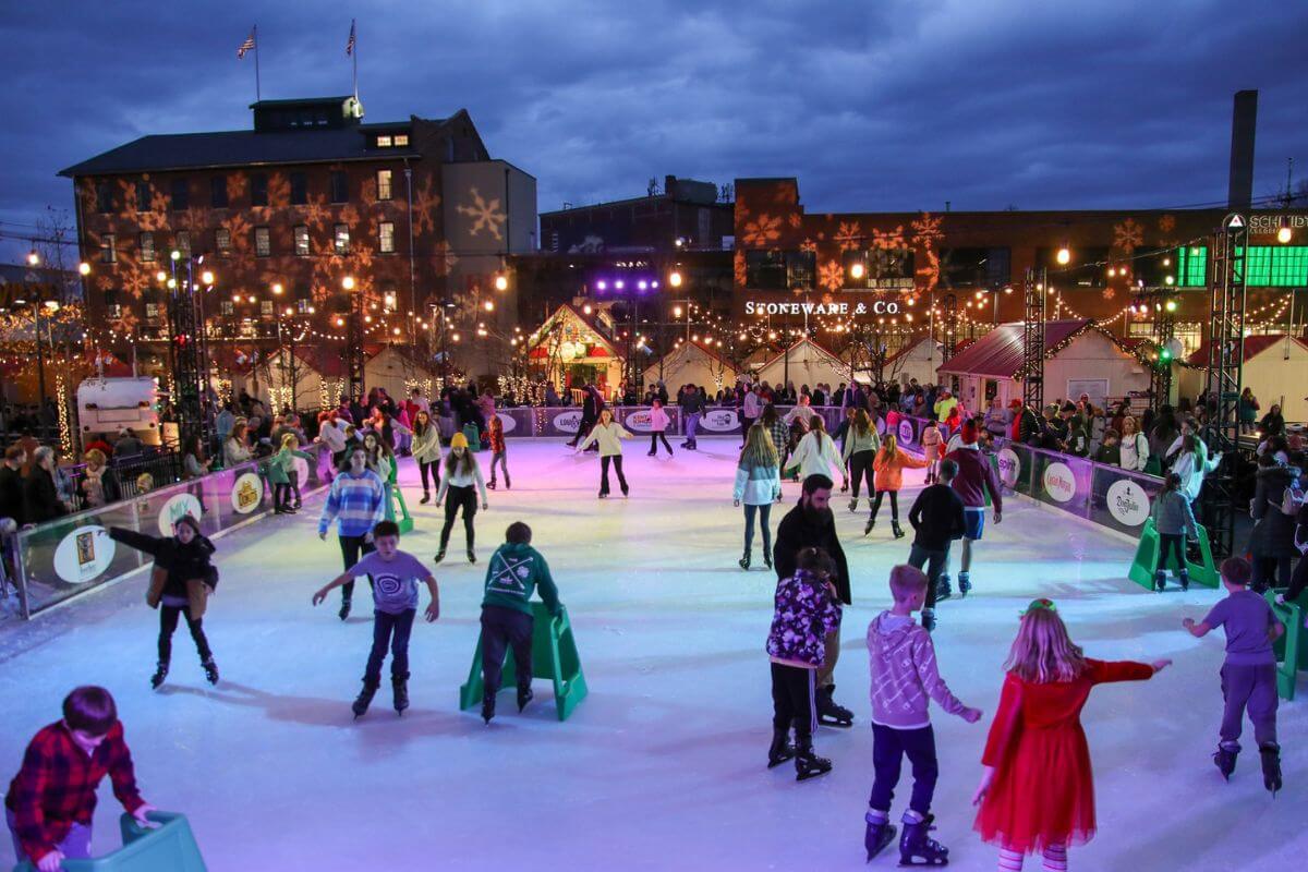 People of various ages ice skating at an outdoor rink in the evening, with festive lights and buildings in the background, enjoying one of the popular January events Louisville has to offer.
