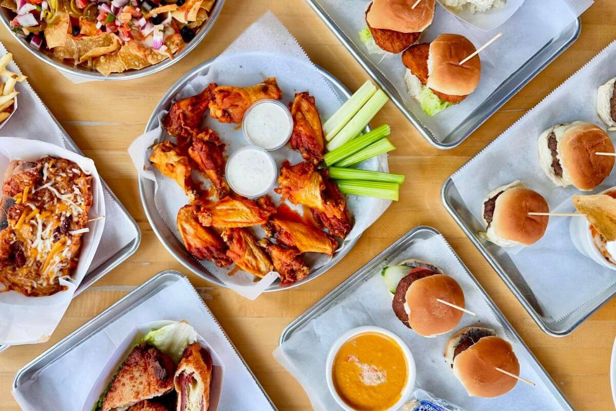 Plates with chicken wings, sliders, nachos, sandwiches, celery sticks, dipping sauces, and soup are arranged on a wooden table—perfect for those searching for Memphis Restaurants Open on New Year's Day.