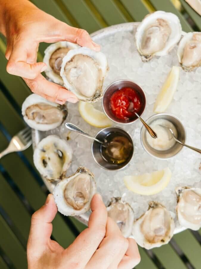 13 Nashville Restaurants OPEN on Christmas Day - 3 A person holds a half-shell oyster over a tray of oysters on ice with lemon wedges and three metal cups of sauces, perfect for those seeking fresh seafood at restaurants open on Christmas Day.