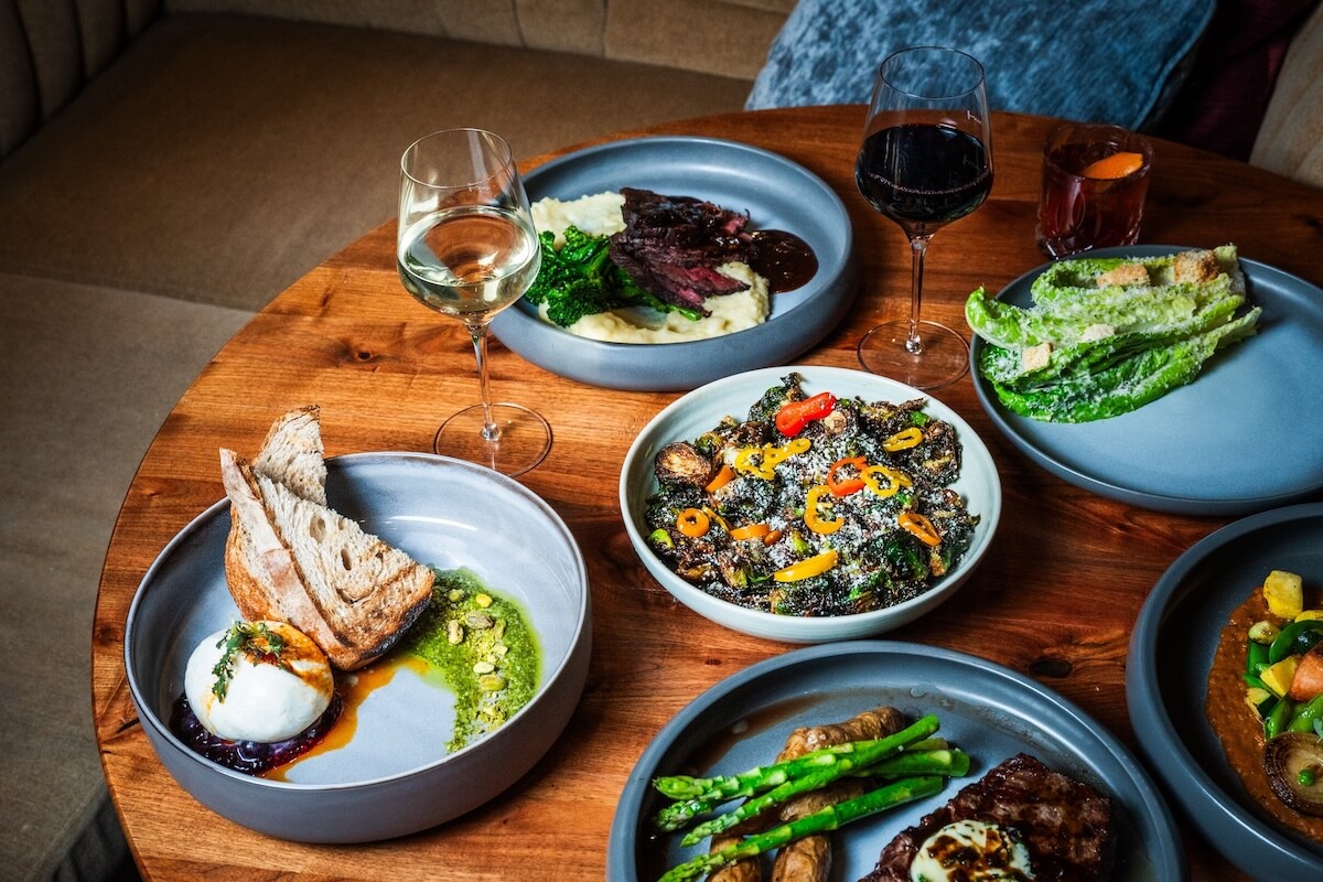 A wooden table set with plated dishes—salad, bread with burrata, steak and mashed potatoes, fresh vegetables, and three glasses of wine—showcases the festive dining found at restaurants open on Christmas Day.