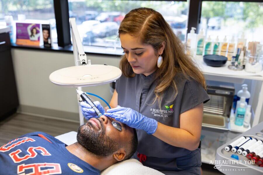 Gift These Memphis Experiences (No Wrapping Required!) - 8 An esthetician wearing gloves performs a facial treatment on a man lying down in a spa clinic, offering one of the unique Memphis gift experiences. Various skincare products and equipment are visible in the background.