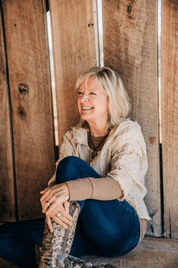 Susan Binkley, with her blonde hair cascading over a light sweater and jeans, sits against the wooden wall, smiling warmly with her legs crossed.