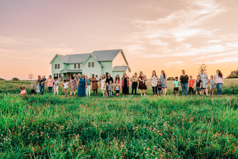 A large group of people stands in front of a green house set against the backdrop of a serene sunset sky.