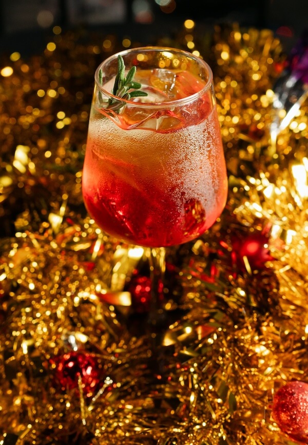 30+ Nashville Holiday Pop-Ups to Get You in the Spirit! - 11 A glass of iced, amber-colored cocktail garnished with a sprig of rosemary sits among gold tinsel and red ornaments, capturing the festive spirit found at Nashville pop up bars.