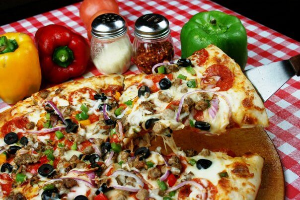 A slice of pizza with various toppings is being served, capturing the essence of Nashville's vibrant culinary scene. Bell peppers, a tomato, and seasoning shakers rest on a checkered tablecloth, evoking nostalgia amid recent restaurant closings.