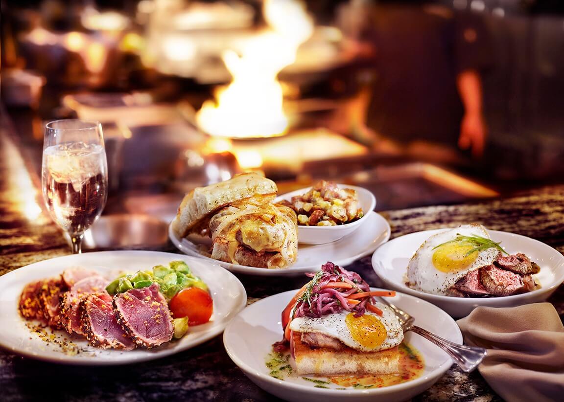 A table featuring gourmet delights: seared tuna, steak with egg, and a sandwich with fries alongside a glass of water. In the background, a chef is busy creating magic. It's a taste of Nashville's culinary scene at its finest.