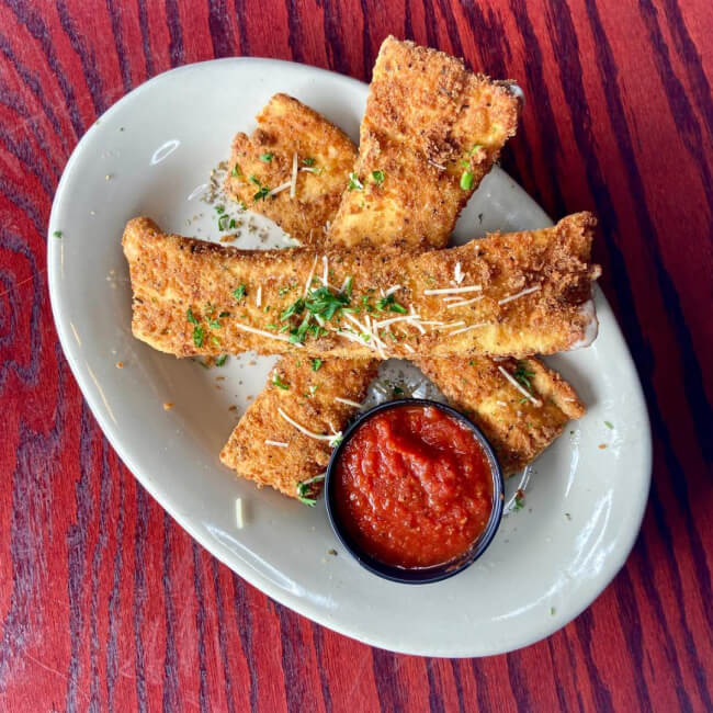 Plate of breaded, fried mozzarella sticks garnished with herbs and grated cheese, served with a small cup of marinara sauce on a white oval plate over a wood-textured table—an appetizer reminiscent of Nashville restaurants' culinary flair.
