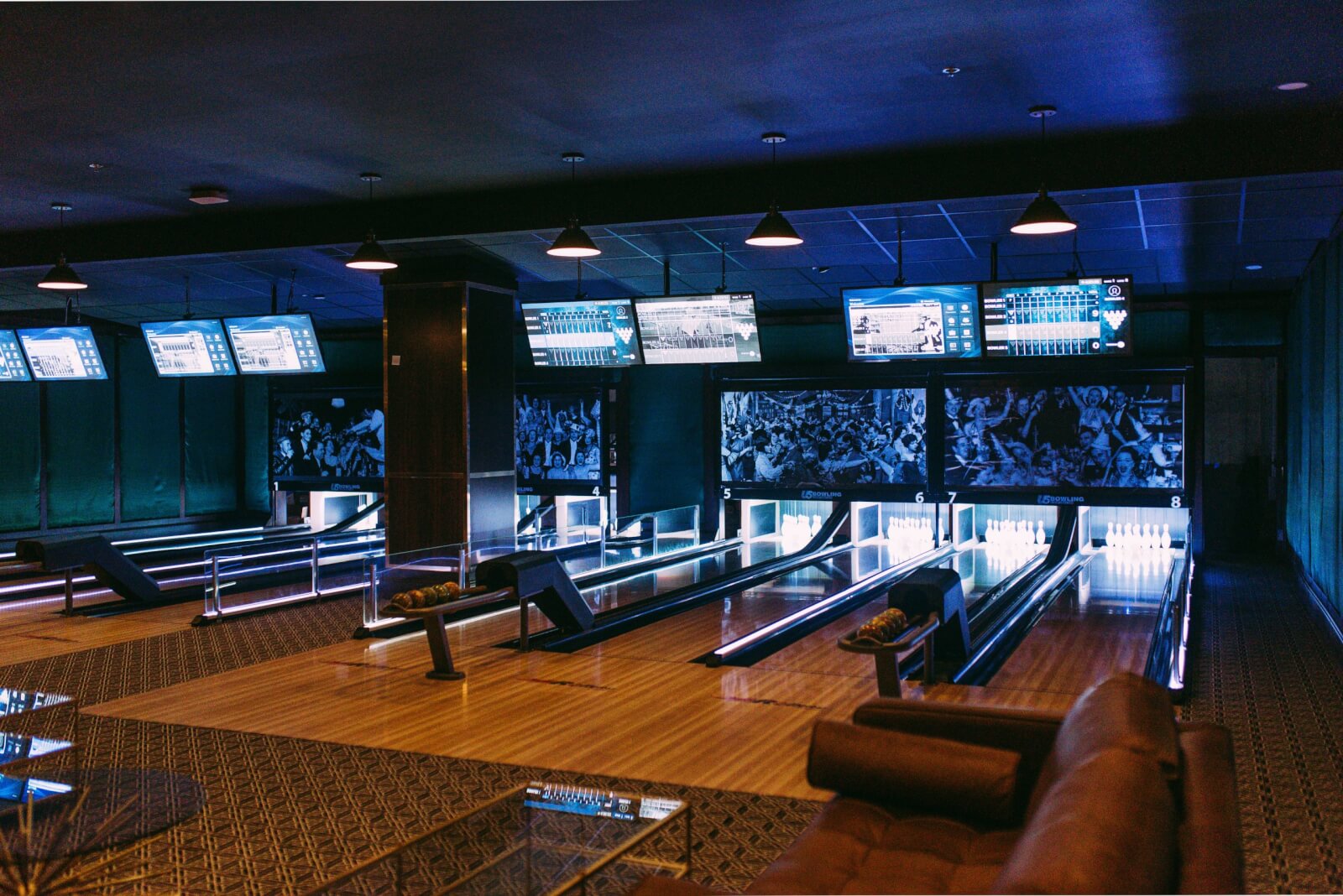 The duckpin bowling alley render at The Lodge features four lanes illuminated by blue lighting. Overhead screens display scores, while a cozy seating area with a sofa invites relaxation in the foreground.