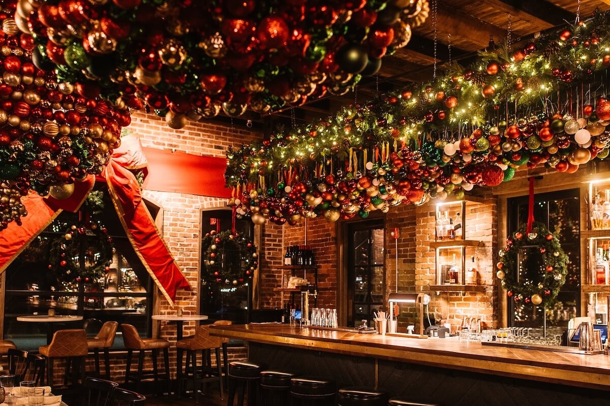 A bar decorated for Christmas with numerous colorful ornaments and garlands hanging from the ceiling, festive lights, and a large red bow on the brick wall—perfect for fans of Nashville pop up bars seeking holiday cheer.
