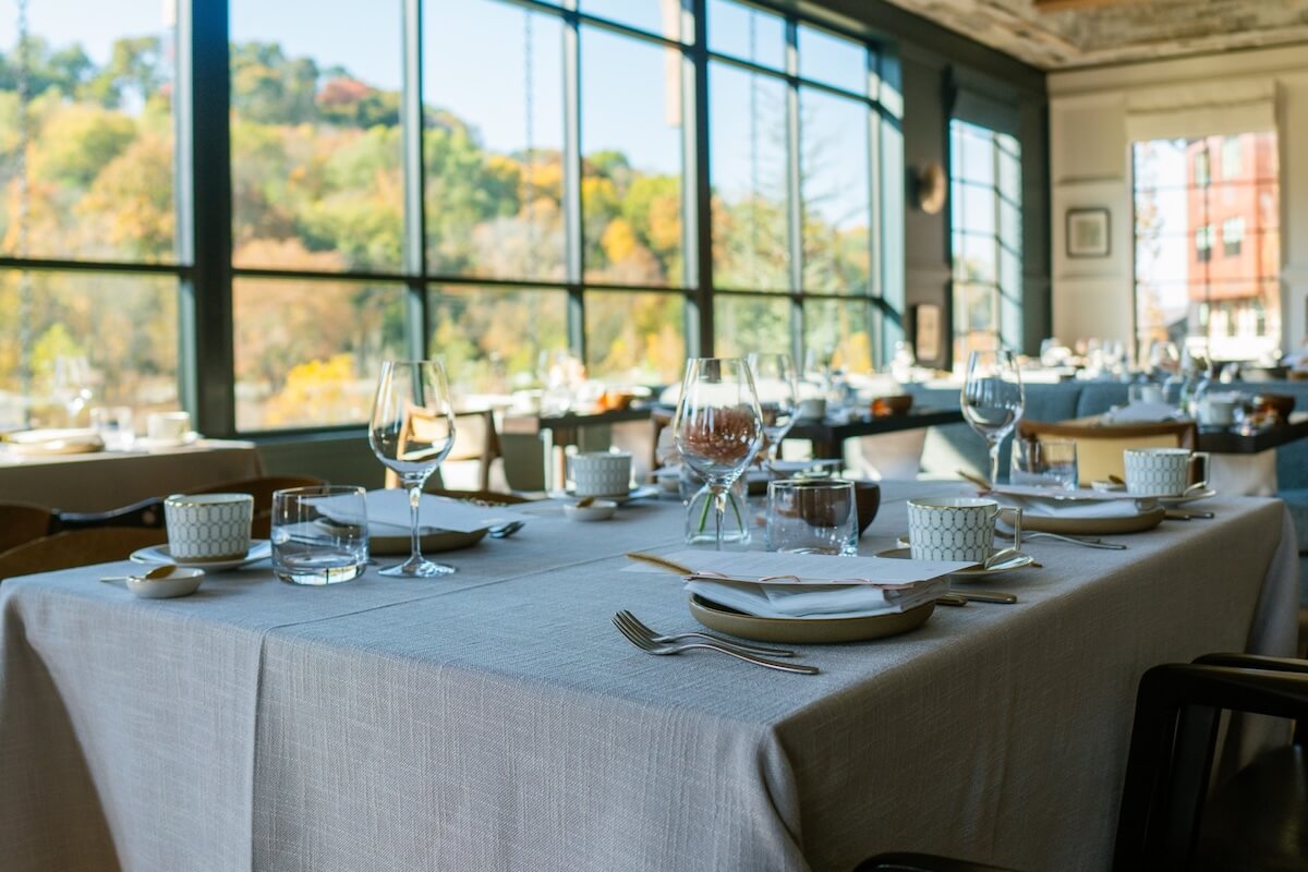 A sunlit restaurant dining area with tables set with glassware, plates, and cups, offers a serene view of trees outside—perfect for enjoying one of the Nashville gift experiences.