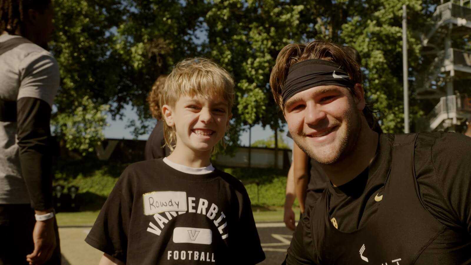 A young boy in a Vanderbilt football t-shirt stands next to a smiling man in athletic wear and a headband, likely his coach at one of the popular Nashville summer camps. Trees and blurred people form the background, capturing a vibrant scene of outdoor adventure and camaraderie.