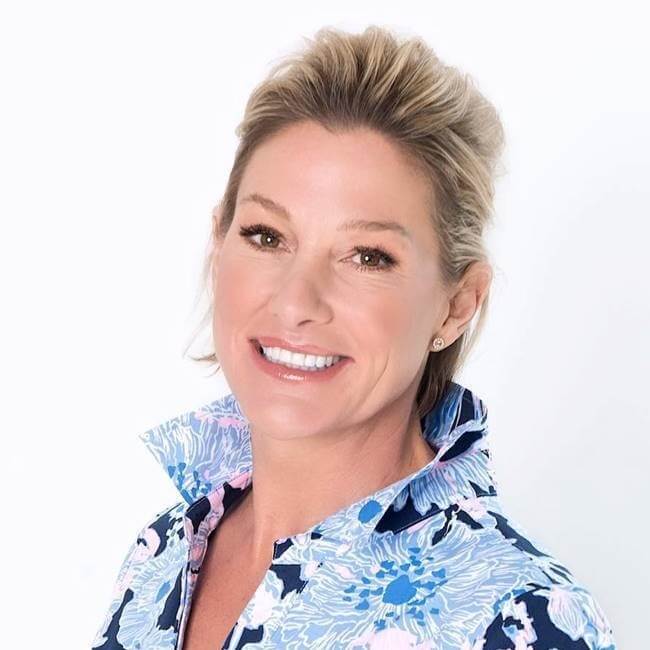 A smiling woman with short, styled hair, standing against a white background, wears a blue floral shirt with the collar up. Her style exudes the charm you'd expect to find on Robertson Academy Rd in Nashville.