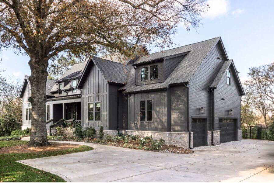 Nestled in the vibrant locale near Robertson Academy in Nashville, TN, this modern gray house features an A-frame roof and a large tree gracing the front yard. The two-car garage and concrete driveway, embraced by low plants, create a picturesque scene under the sunny blue sky.