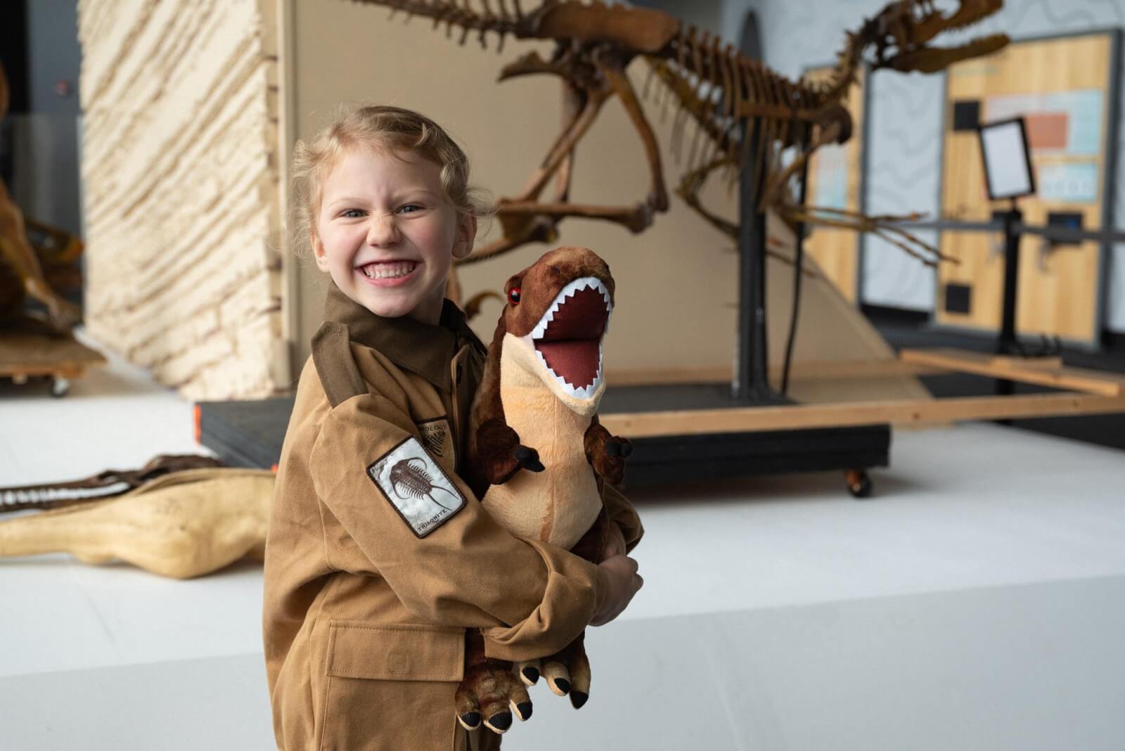 A child beams with joy, clutching a toy dinosaur while standing before a towering skeleton display, dreaming of adventures at Nashville summer camps.