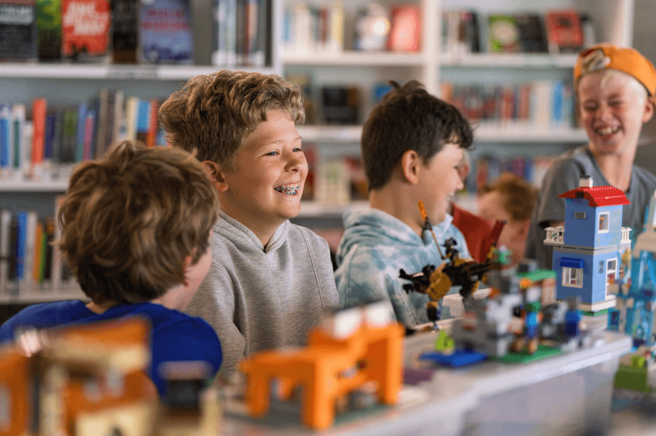 A group of children are gathered around a table with toys and blocks, smiling and chatting in a brightly lit room with shelves of books in the background, reminiscent of the joyful atmosphere at Nashville summer camps.