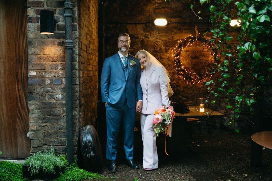 Nashville’s 10 Most Popular Articles of 2025 - 5 In an intimate Edinburgh wedding setting, a bride and groom stand against rustic brick walls adorned with greenery. The bride holds a bouquet of flowers. They are smiling and dressed in formal attire.