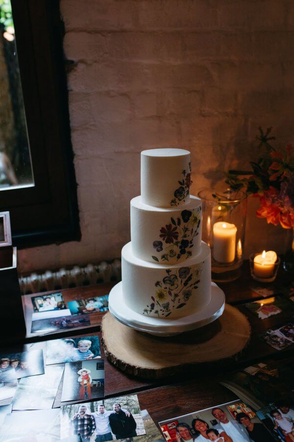 A three-tiered wedding cake with floral designs is beautifully displayed on a wooden table surrounded by intimate photographs, candles, and a delicate floral arrangement, perfectly capturing the romantic spirit of an Edinburgh celebration.
