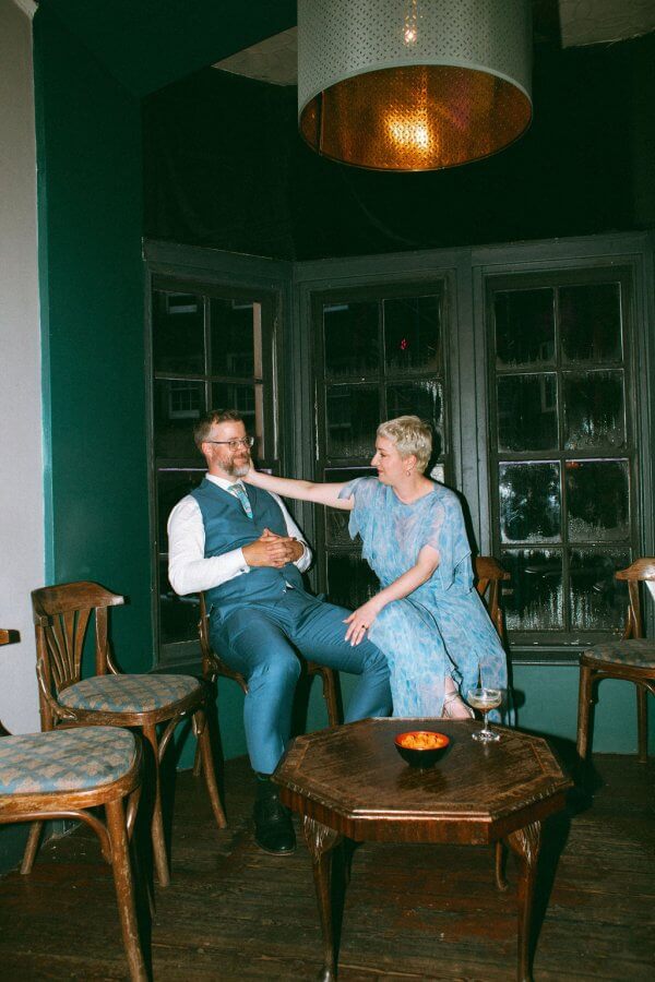 In an intimate setting, a person in a blue dress sits with a person in a blue suit, smiling and talking in a dimly lit room. Wooden chairs surround them, and on the table before them rests a bowl and a glass.