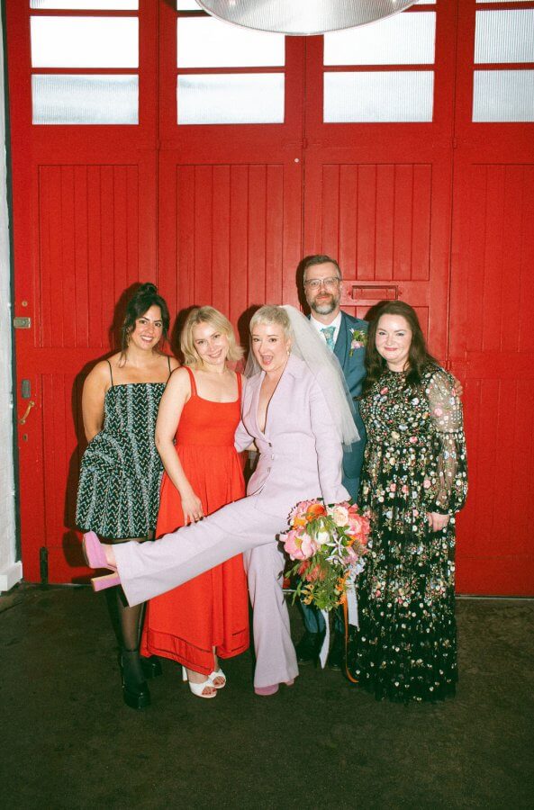 A lively group of five poses in front of red doors, capturing the joy of an Edinburgh wedding; one person in a lavender suit playfully lifts a leg, bouquet in hand.