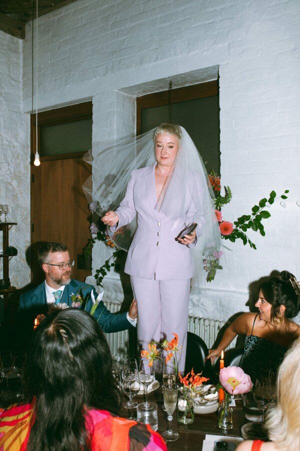 At an intimate Edinburgh wedding, a bride in a lavender suit and veil stands confidently on a table, holding her phone. Guests seated around the table watch her with amused anticipation.
