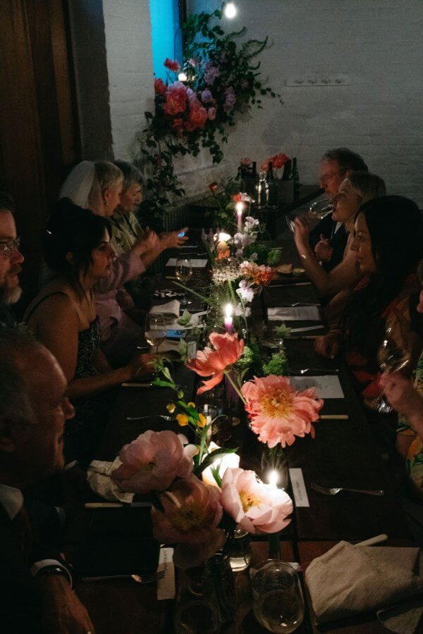 People are sitting around a dinner table adorned with pink flowers and lit candles, engaged in conversation in a dimly lit room, capturing the essence of an intimate wedding.