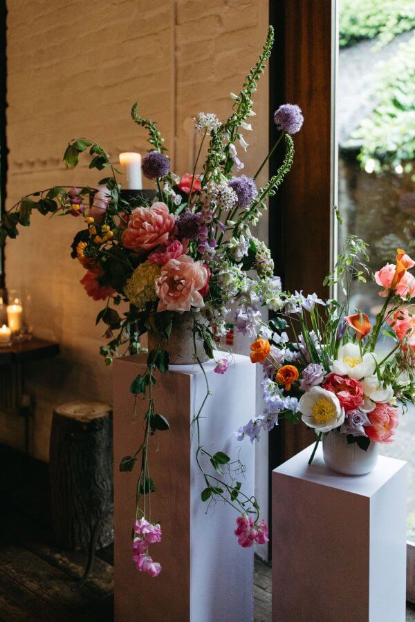 Elegant floral arrangements with pink, purple, and orange flowers grace white pedestals near a window, setting the stage for an intimate Edinburgh wedding. Candles and rustic decor add a vibey ambiance in the background.