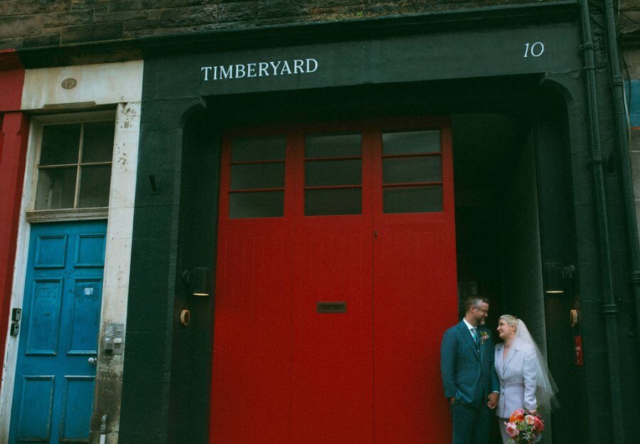 An intimate moment unfolds as the bride and groom stand outside a red door under the "Timberyard" sign, capturing the charm of an Edinburgh wedding.