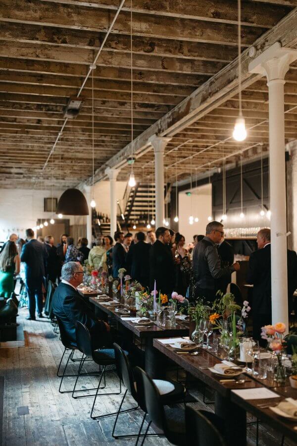A large Edinburgh wedding unfolds in a rustic venue with wooden beams. Guests mingle near long, decorated tables adorned with floral centerpieces and soft overhead lighting, creating an enchanting ambiance.