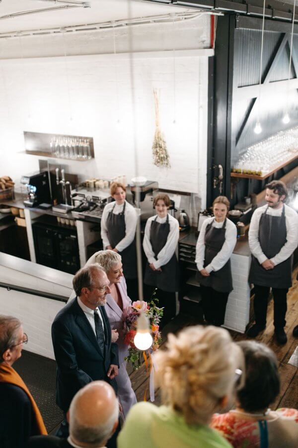 A father and daughter stand together holding flowers in a kitchen setting reminiscent of an elegant Edinburgh wedding, observed by people in the foreground and staff in uniforms in the background.