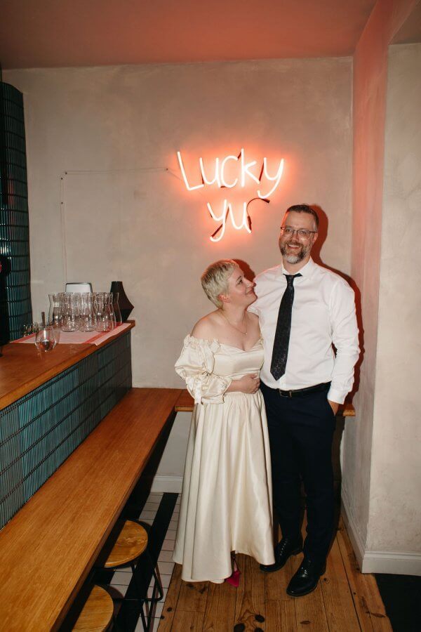 A couple stands in a bar, smiling beneath a "Lucky" neon sign on the wall. She wears an off-shoulder dress; he dons a white shirt and tie. Celebrating their Edinburgh wedding, they embrace the joy of their special day in style and charm.
