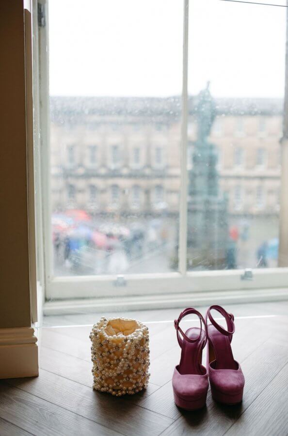 Pink high heels and a beaded decorative object rest on a wooden floor by a rainy window, framing blurred Edinburgh buildings outside, setting the perfect scene for an enchanting wedding day.