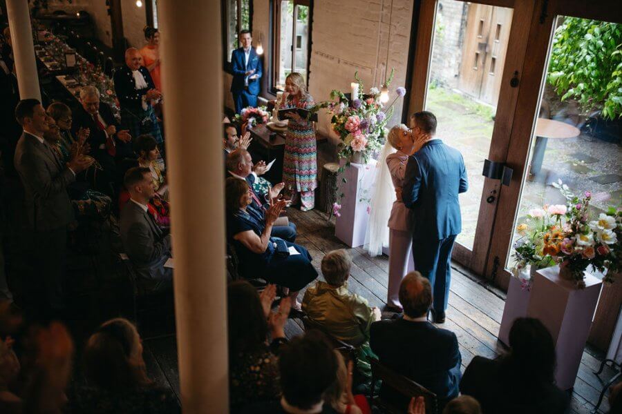 At an intimate wedding in a rustic venue, a couple kisses as guests watch, surrounded by enchanting floral arrangements and gentle natural light. The whole scene embodies the perfect vibey wedding atmosphere.