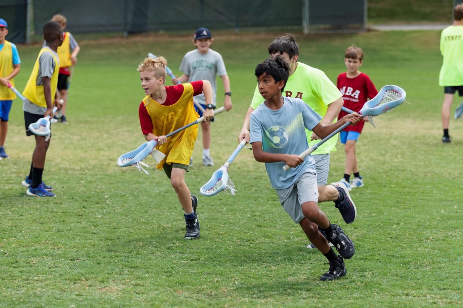 Children enjoying lacrosse at a Nashville summer camp on a grassy field, wearing casual sportswear and holding lacrosse sticks. Some wear yellow and red bibs, while other children and a staff member are visible in the background.