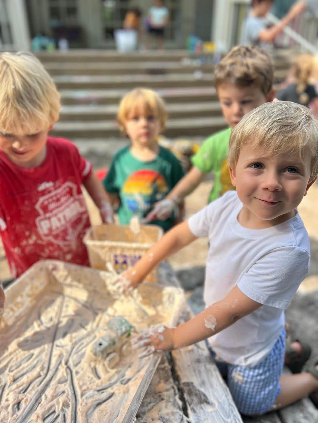 The 2026 Memphis Summer Camp Guide - 4 Four young children play with a sensory bin filled with a white, foamy substance outdoors at one of the Memphis summer camps, using their hands and small objects. Steps and a building appear in the background.