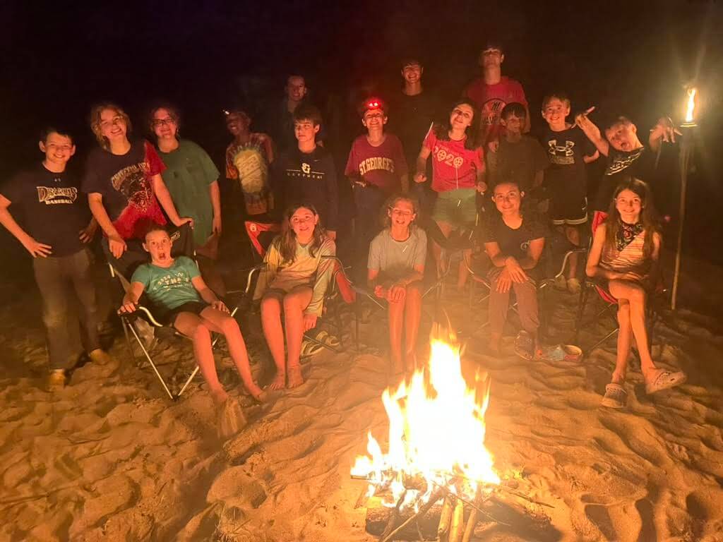The 2026 Memphis Summer Camp Guide - 7 A group of teens and kids sit and stand around a campfire at night on sandy ground, smiling at the camera during one of the fun-filled Memphis summer camps.