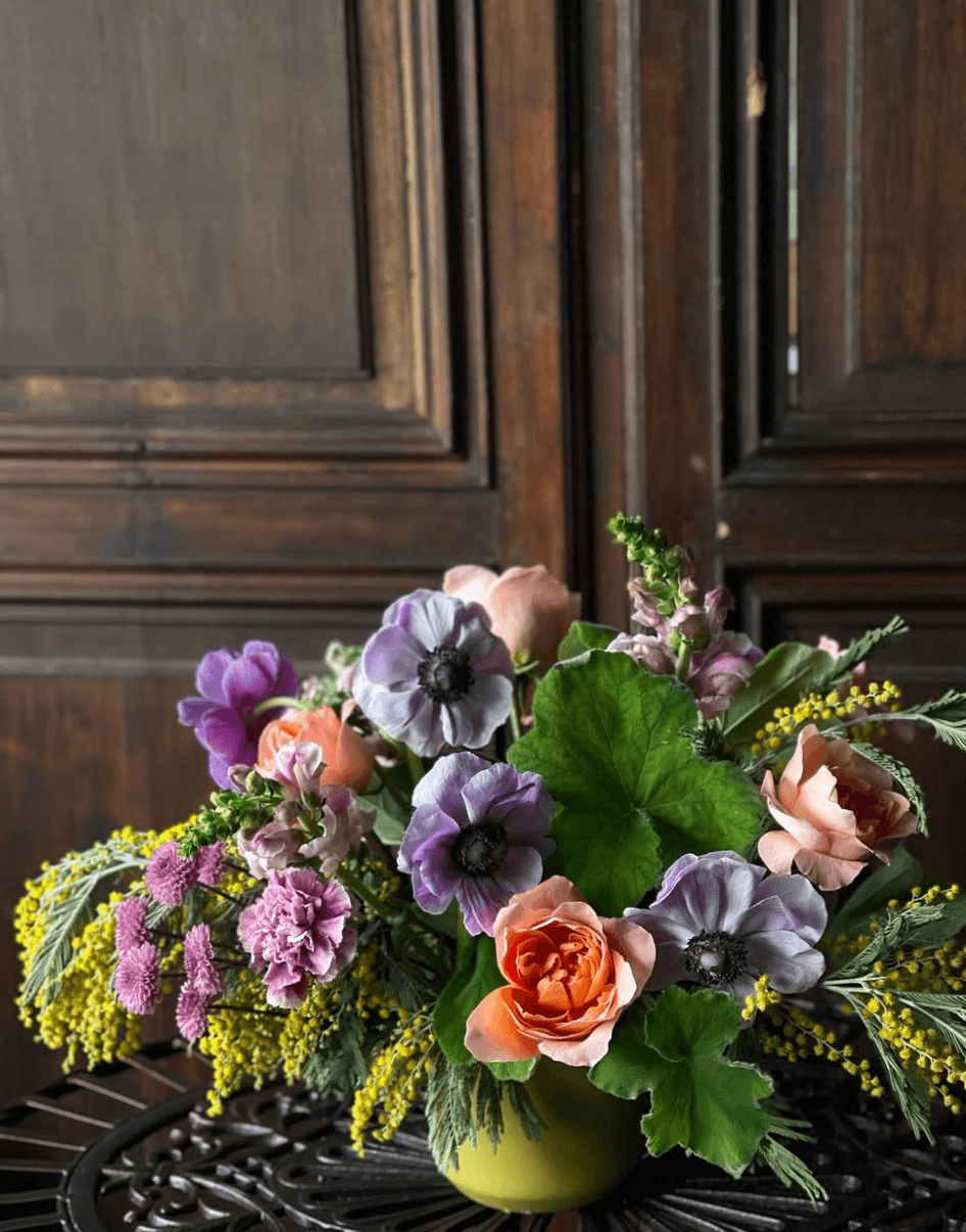 A stunning floral arrangement from Nashville florists graces a green vase. It showcases vibrant orange roses, purple and pink blooms, complemented by lush greenery, all set against a dark wooden backdrop.
