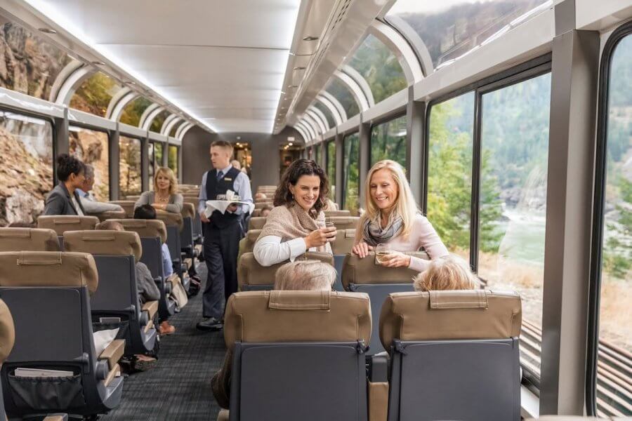 StyleBlueprint's 15 Most Popular Articles of 2025 - 9 Passengers in a scenic train car share drinks, embracing the latest travel trends, with large windows revealing a forested landscape.