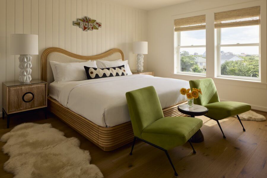23 New Boutique Hotels in the South - 3 A bedroom in one of the new boutique hotels features a large bed, green chairs, and a small table adorned with flowers. Two lamps rest on wooden side tables. The room's white walls are complemented by two windows and fluffy white rugs on the wooden floor.
