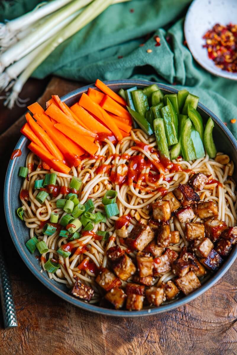 A garlicky noodle bowl topped with diced tofu, sliced carrots, green onion, green bell pepper, and a drizzle of sauce. Scallions and red pepper flakes are nearby.