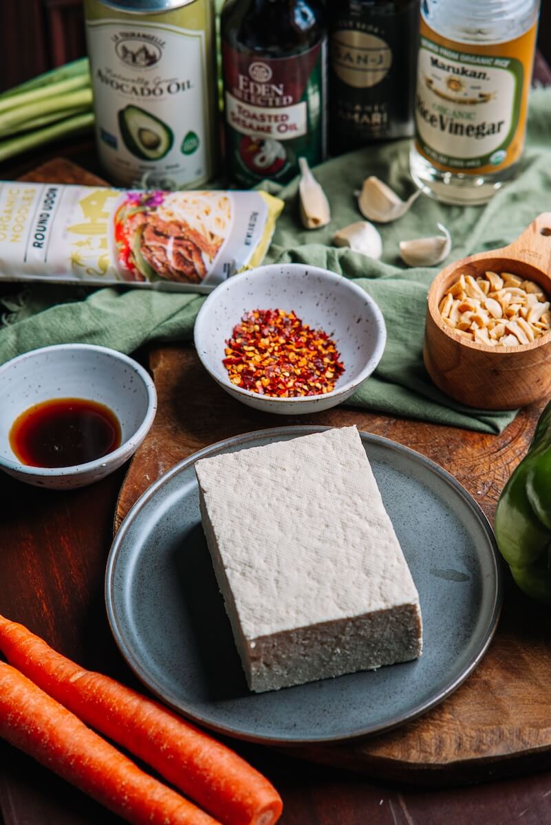 A block of garlicky baked tofu sits on a plate, surrounded by carrots, chili flakes, soy sauce, oils, nuts, garlic cloves, and vinegar packaging on a wooden surface. Perfect for tossing into an Asian noodle bowl.