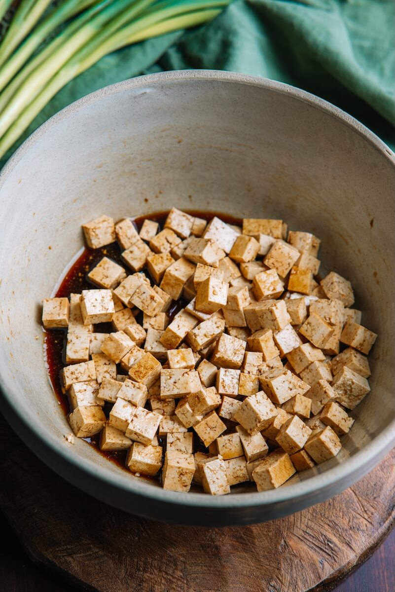 Diced tofu marinating in soy sauce sits in a bowl, ready to complement garlicky noodles. Surrounded by fresh green onion stalks and a vibrant green cloth, this scene embodies the essence of an Asian Noodle Bowl feast.