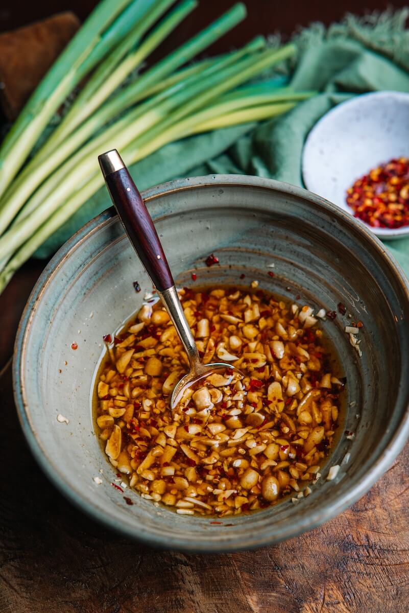 A bowl of garlicky chili oil with peanuts and a spoon inside, set on a wooden surface. Green onions and a small dish with crushed red pepper are in the background, perfect for drizzling over an Asian noodle bowl.