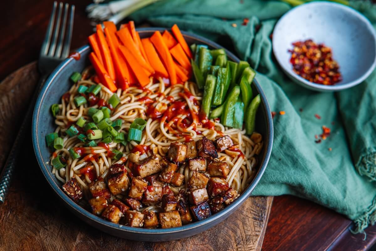 A fragrant Asian Noodle Bowl featuring garlicky baked tofu, sliced carrots, green peppers, and scallions drizzled with chili sauce. A fork waits ready for a delicious bite as a small bowl of fiery chili flakes sits in the background.