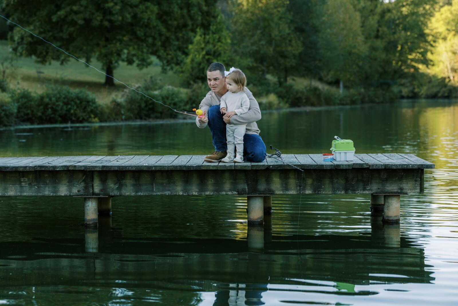 A man teaching a toddler to fish off a dock over the water.