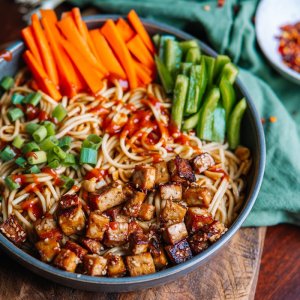 A bowl of noodles topped with diced tofu, sliced carrots, bell peppers, green onions, and a drizzle of sauce, placed on a wooden surface with a fork and a small dish nearby.