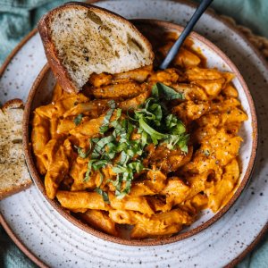 A bowl of pasta with creamy orange sauce, topped with chopped basil. Two slices of garlic bread are placed on the side.