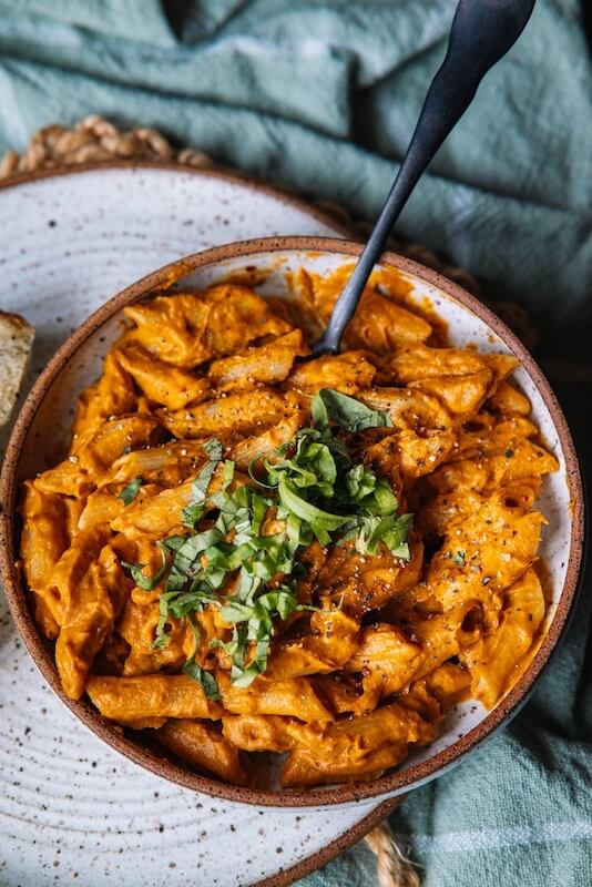 A bowl of creamy penne pasta drizzled with a rich pasta sauce, topped with fresh basil and ground pepper, with a fork resting inside.