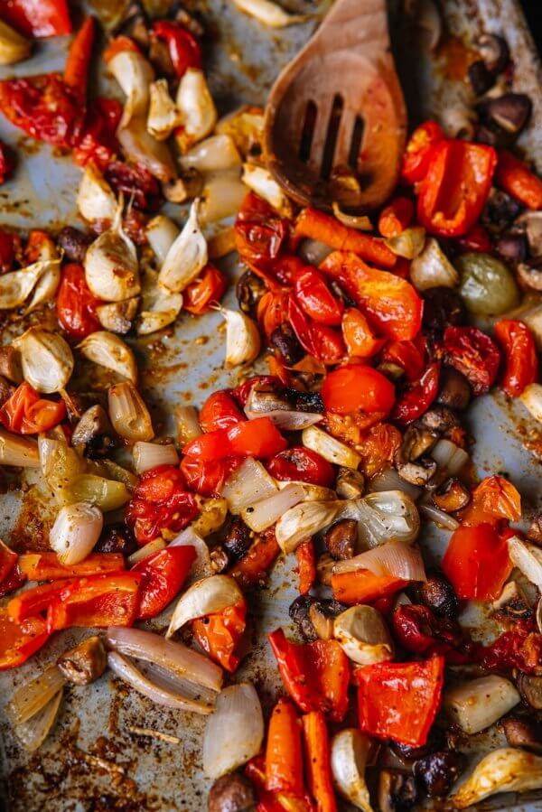 A baking tray filled with roasted red bell peppers, onions, garlic, and mushrooms, perfect for stirring into a vibrant pasta sauce with a wooden spoon.