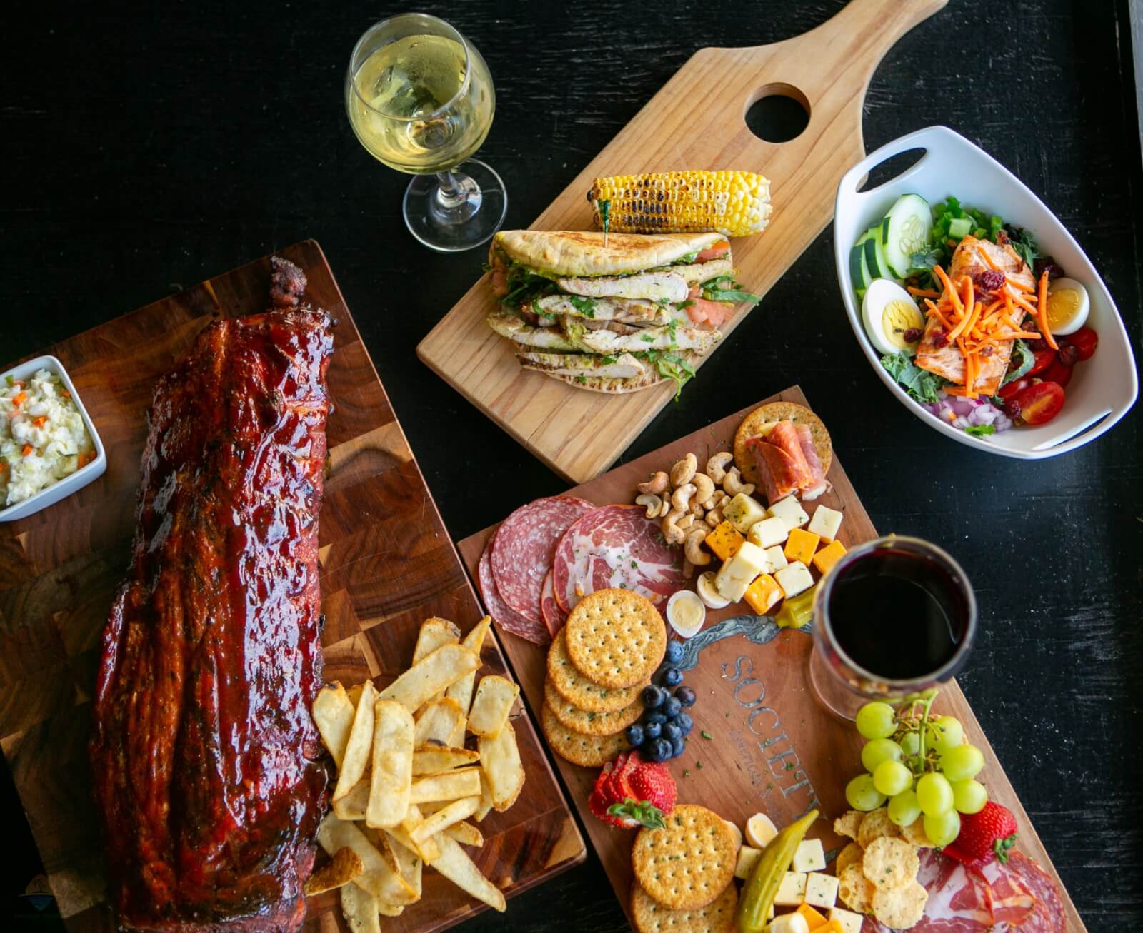 A table set for a feast during Tennessee vacations, featuring a platter of ribs, fries, and coleslaw, a charcuterie board, a glass of wine, a salad, and panini with corn.